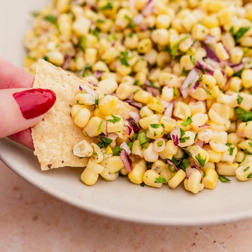 A person is dipping a tortilla into a bowl of corn salsa.