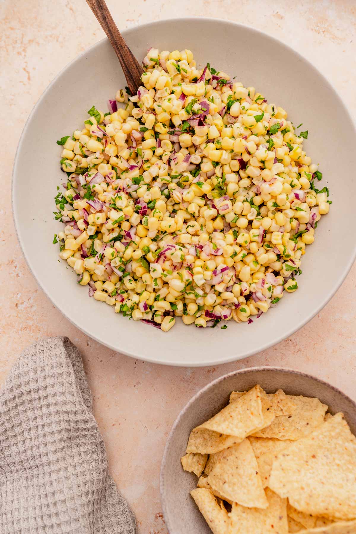 A bowl of chipotle corn salad with tortilla chips and a wooden spoon.