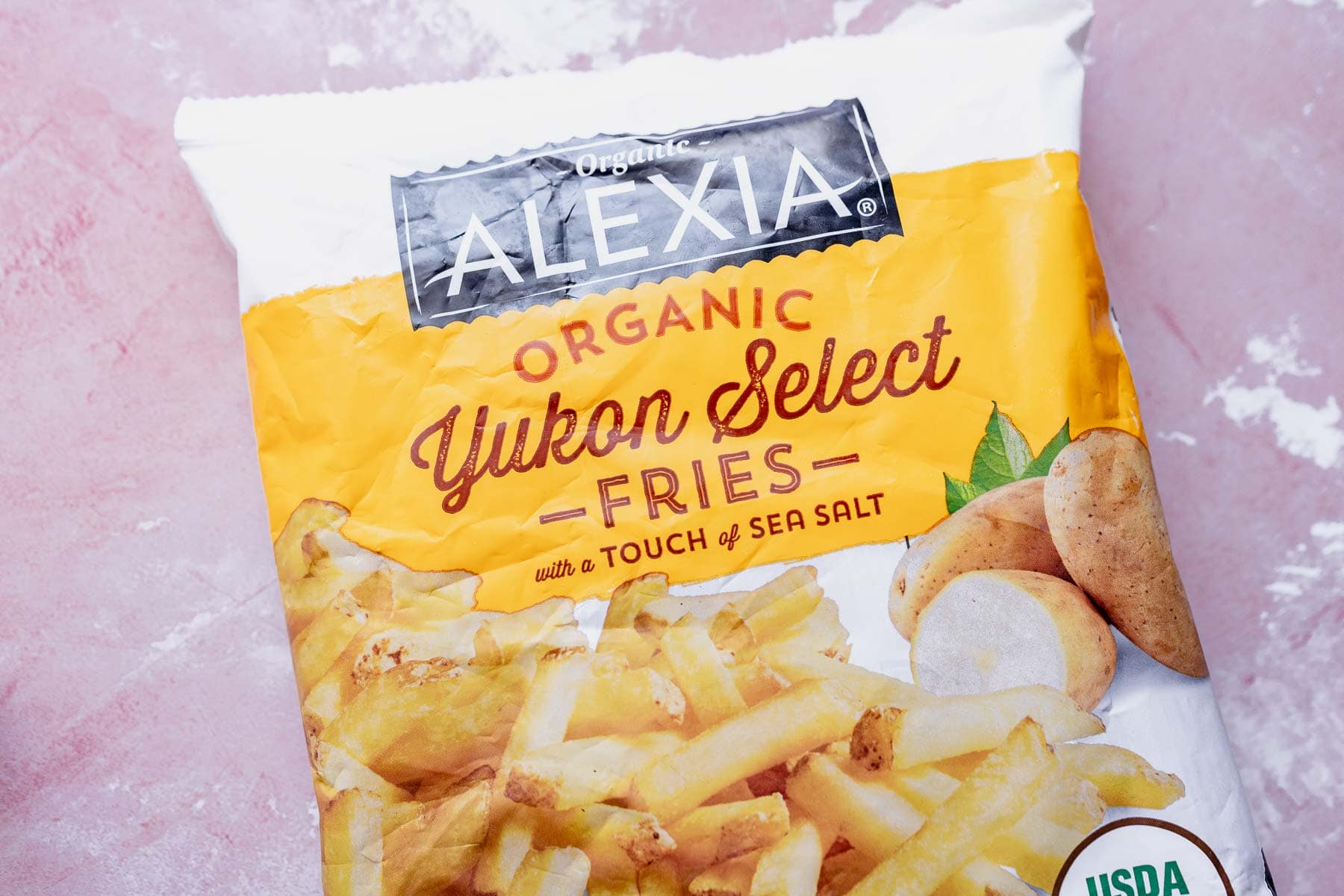 Bag of alexia organic yukon select air fryer frozen french fries on a pink surface.