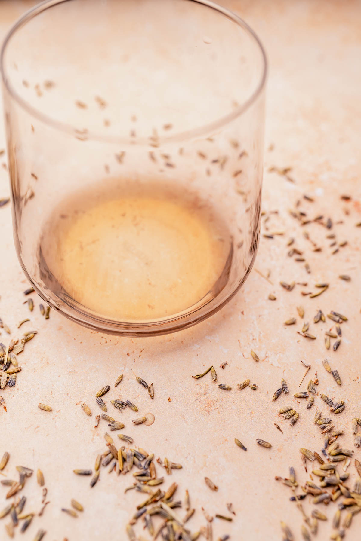 An empty glass with traces of an Iced Lavender Oatmilk Latte, surrounded by scattered cumin seeds on a beige surface.