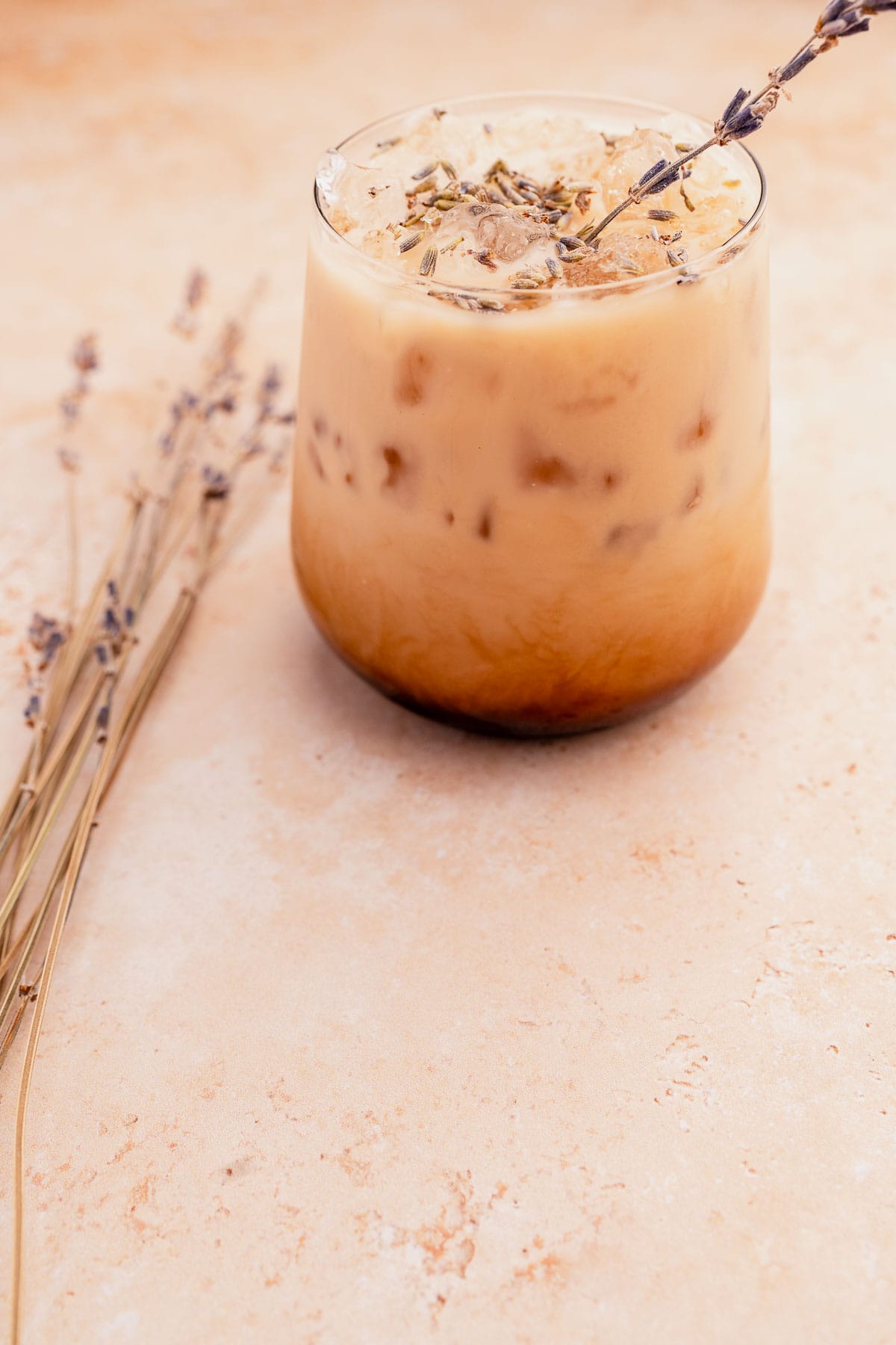 A glass of iced lavender oatmilk latte garnished with lavender on a beige surface, with dry lavender stems beside it.