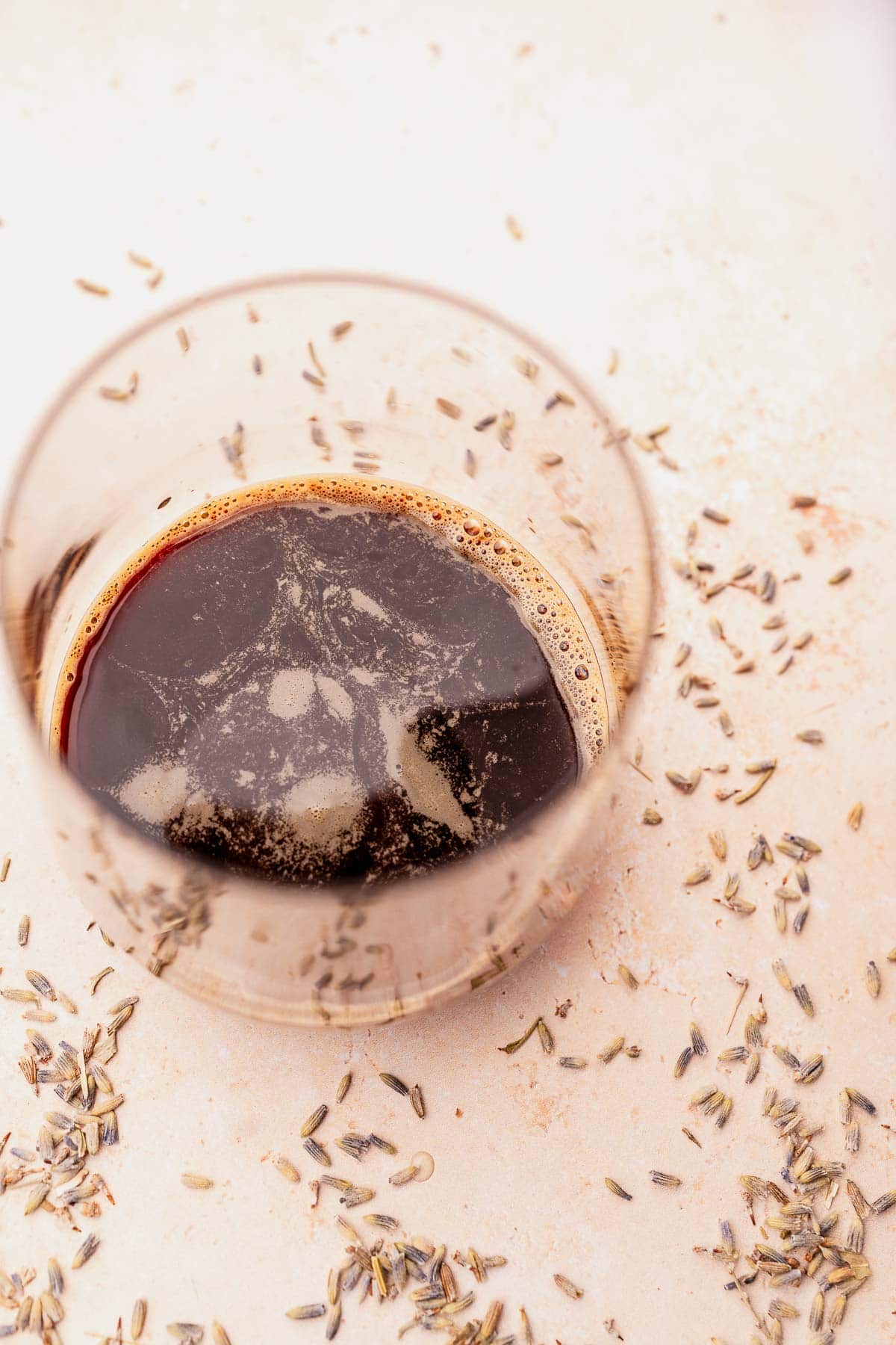 A glass of dark beer with foam on top surrounded by scattered grains on a light surface.