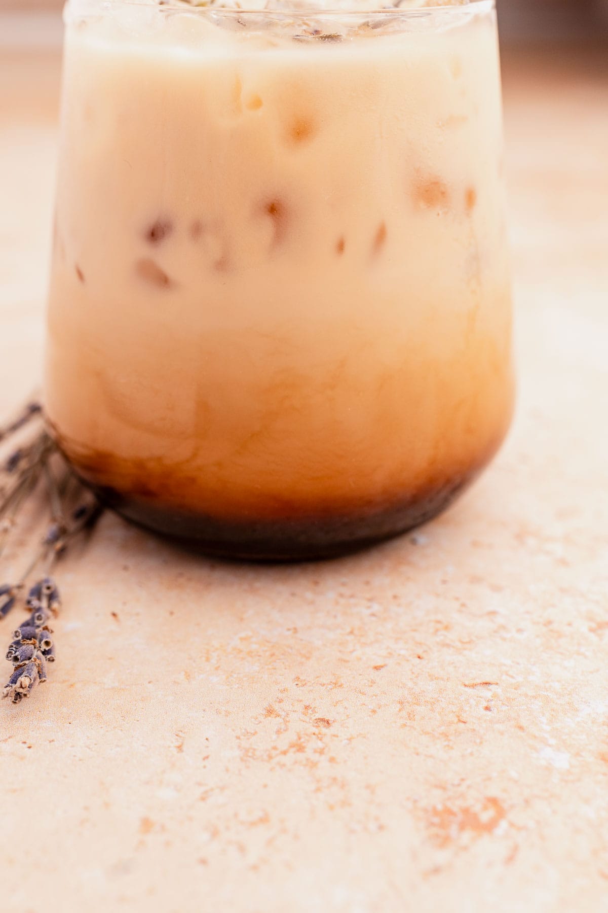 Glass of iced lavender oatmilk latte with layers of milk and espresso, accompanied by lavender sprigs on a textured surface.