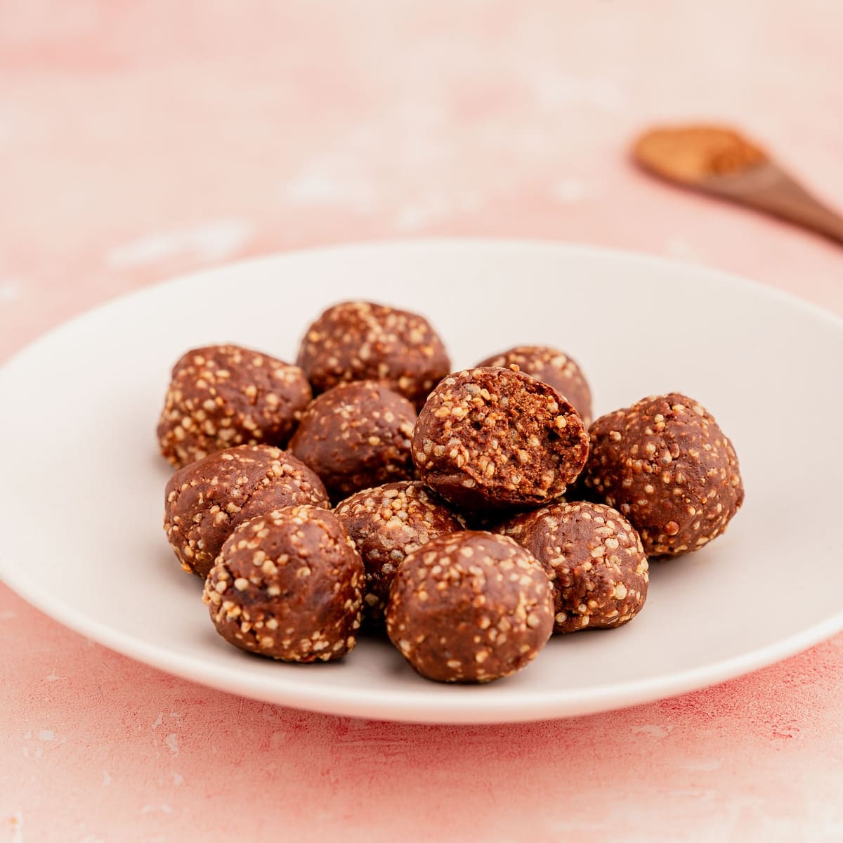 A white plate containing several brown, round quinoa crunch bites on a pink surface, with a wooden spoon in the background.
