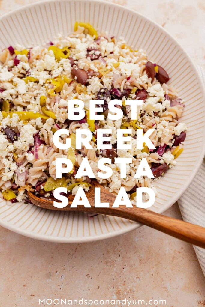 A bowl of Greek pasta salad with feta cheese, olives, and onions, captioned "best Greek pasta salad" on a textured table.