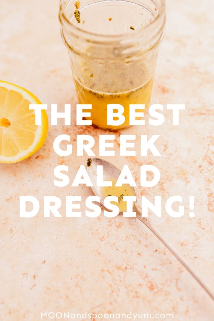 A jar of Greek salad dressing with a lemon half and a spoon, labeled "the best Greek salad dressing!" on a textured surface.