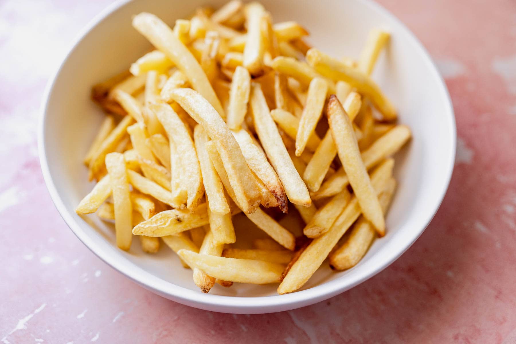 A bowl of golden-brown air fryer frozen french fries on a pink surface.