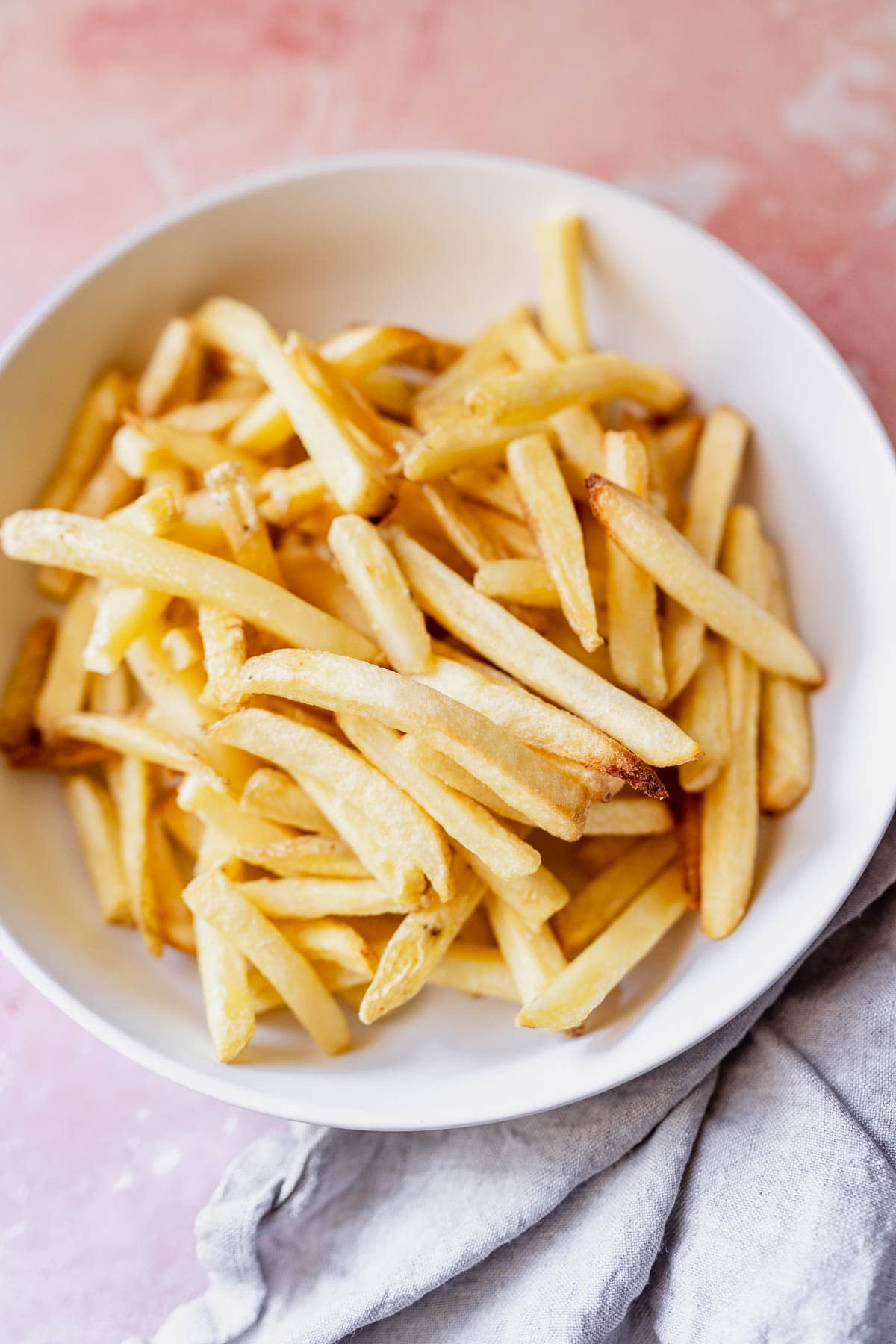 A bowl of golden air fryer frozen French fries on a pink surface with a gray napkin.