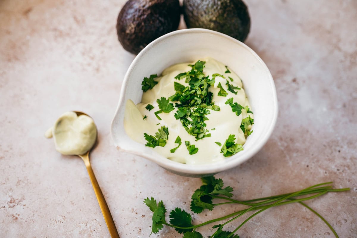 Bowl of creamy avocado sauce garnished with chopped cilantro, alongside whole avocados and a spoonful of sauce on a marble surface.