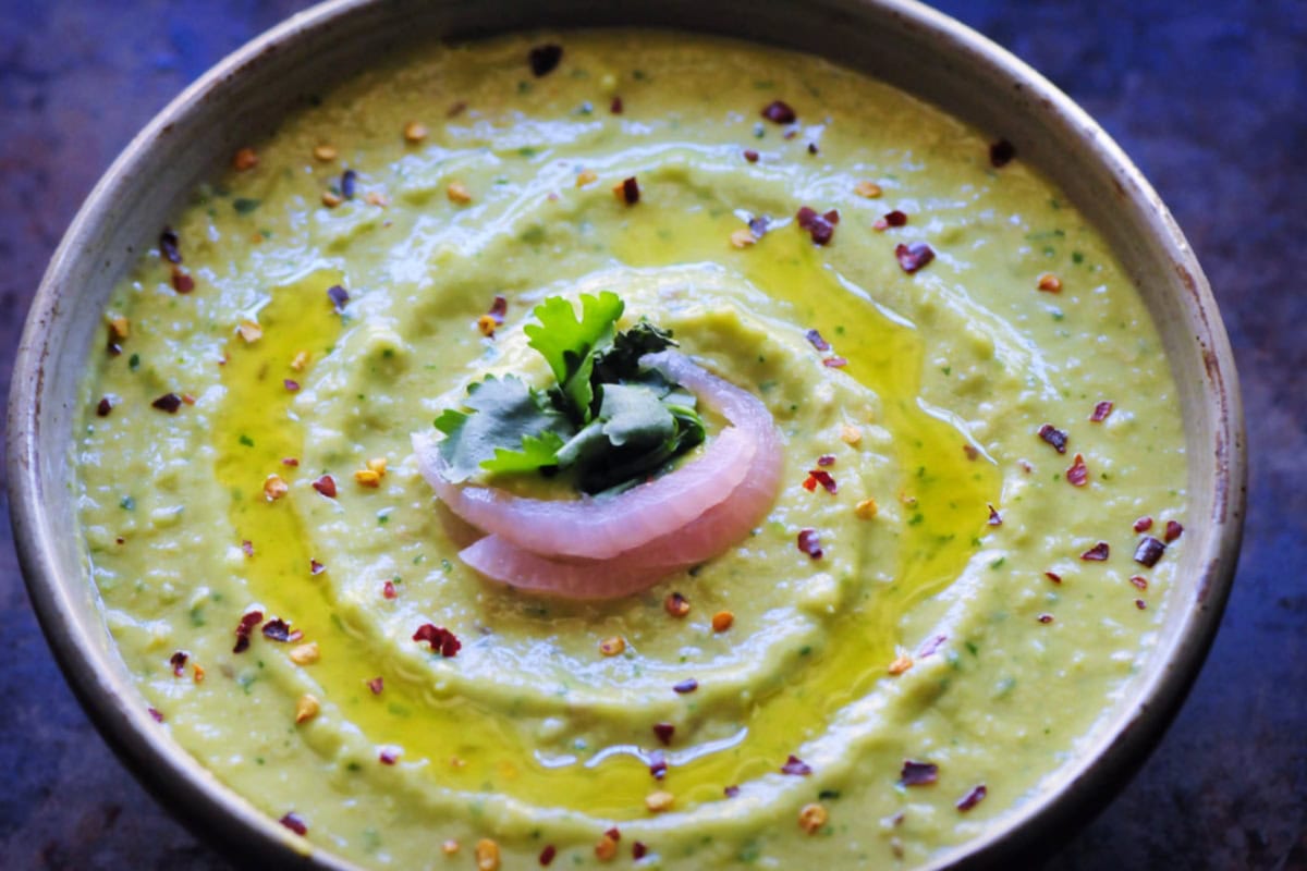 A bowl of creamy green soup garnished with cilantro, onion, and red pepper flakes, served in a rustic bowl on a dark surface.