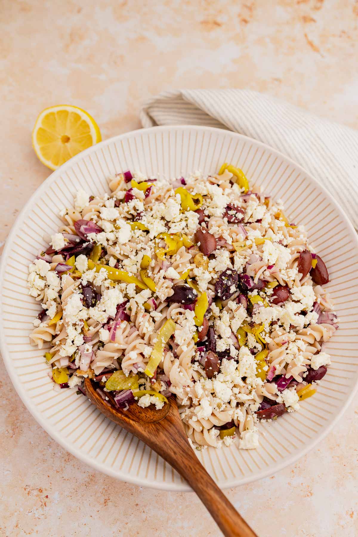 Bowl of Greek orzo pasta salad with feta cheese, olives, and diced vegetables, accompanied by a wooden spoon and a lemon wedge on a marbled countertop.