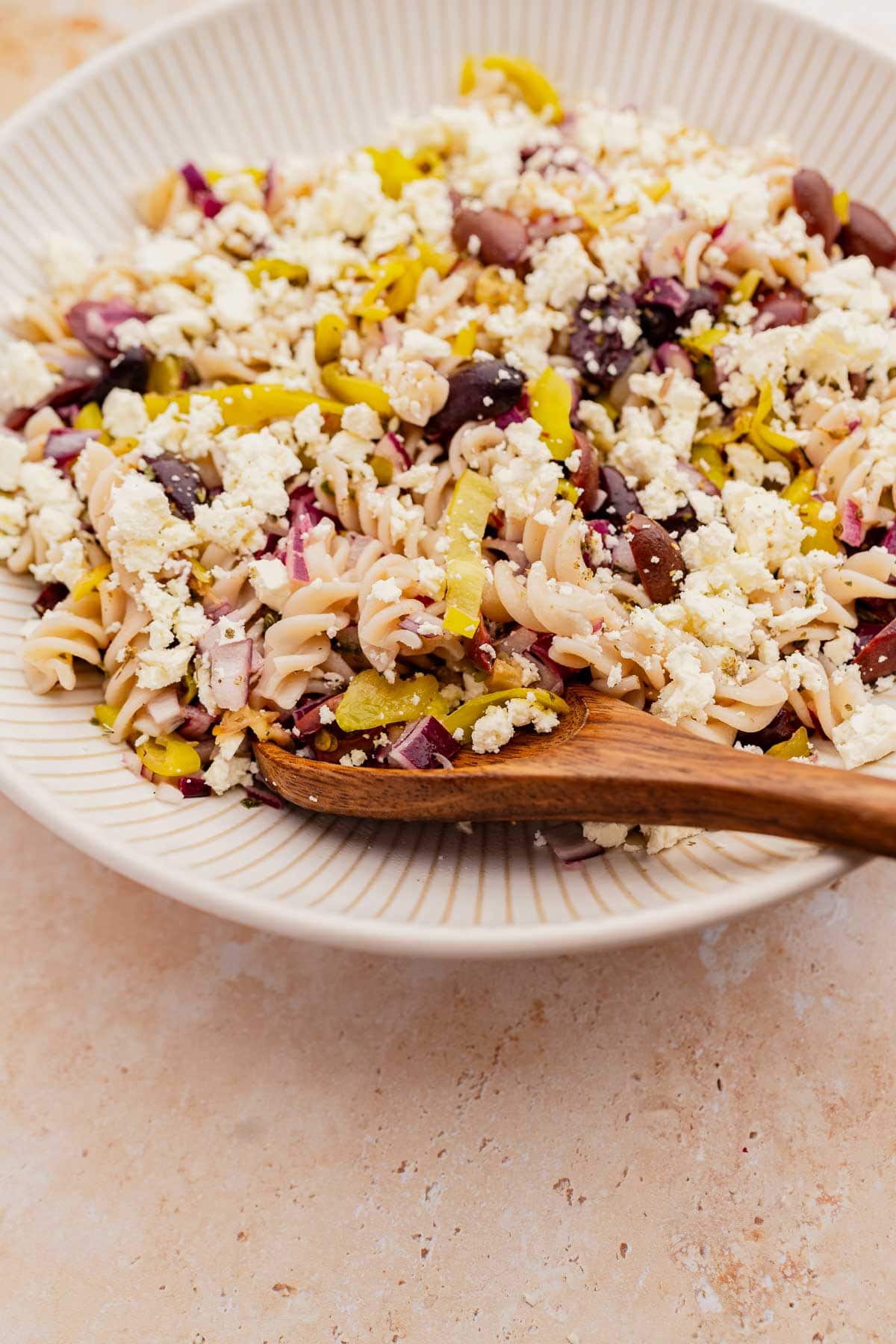 A bowl of Greek pasta salad with olives, feta cheese, and red onions, mixed together with a wooden spoon.