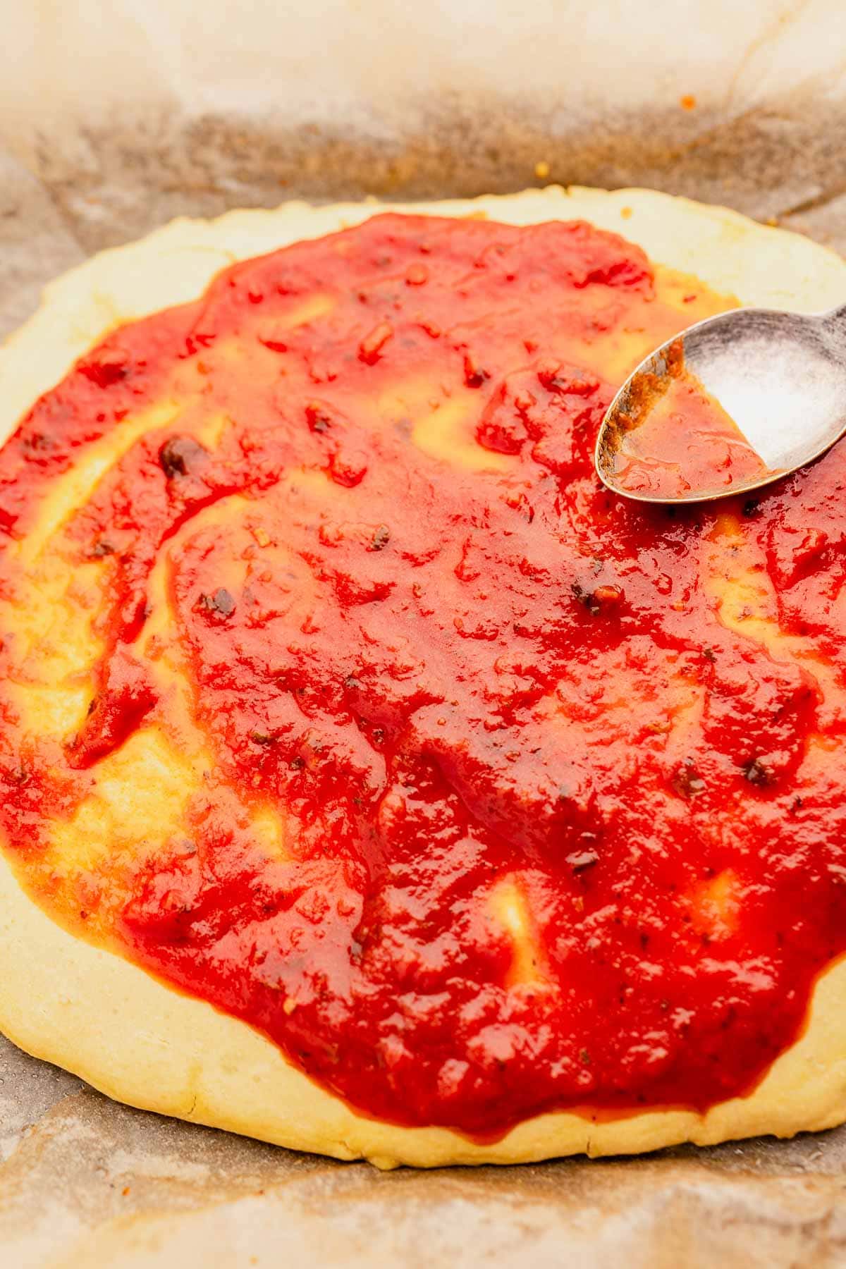 A homemade chickpea flour pizza crust topped with tomato sauce and a spoon resting on the surface, on parchment paper.