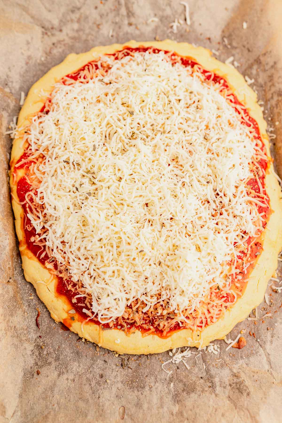 A freshly made chickpea flour pizza crust with a generous topping of shredded cheese and tomato sauce on a baking sheet.