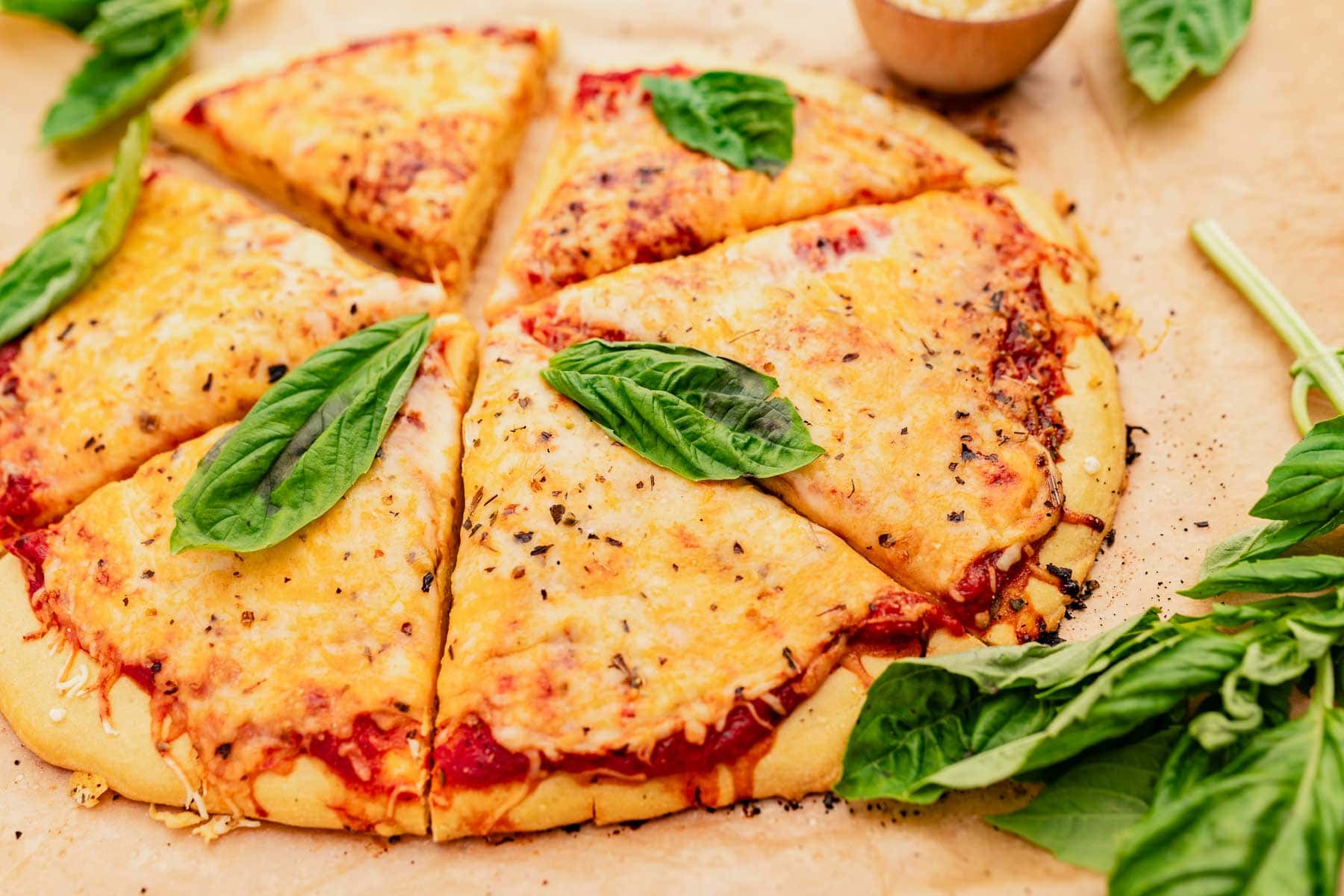 Sliced cheese pizza with a chickpea flour crust garnished with fresh basil leaves on a light surface, with whole basil leaves and a small bowl of sauce nearby.