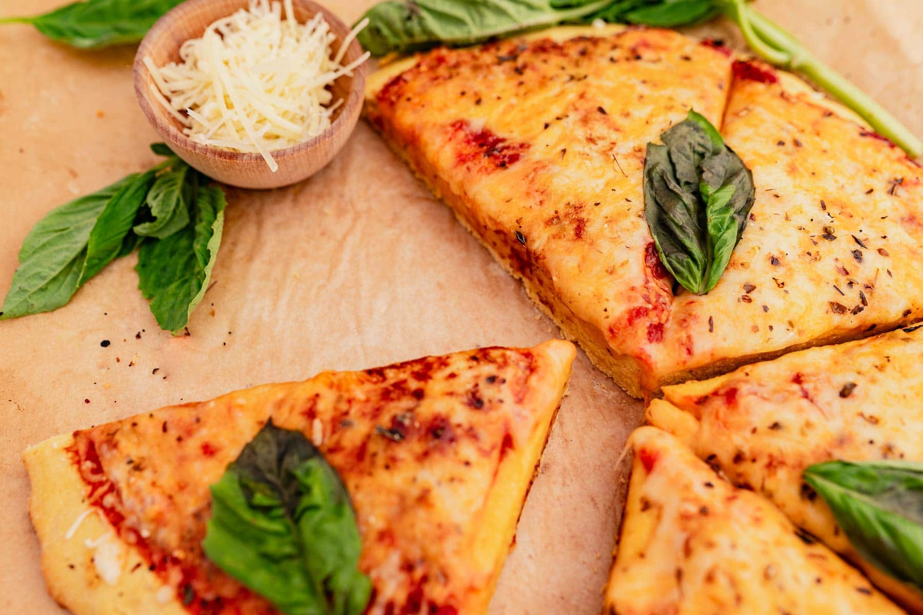 Slices of cheese pizza with chickpea flour crust, fresh basil leaves, and a small bowl of grated cheese on a brown surface.