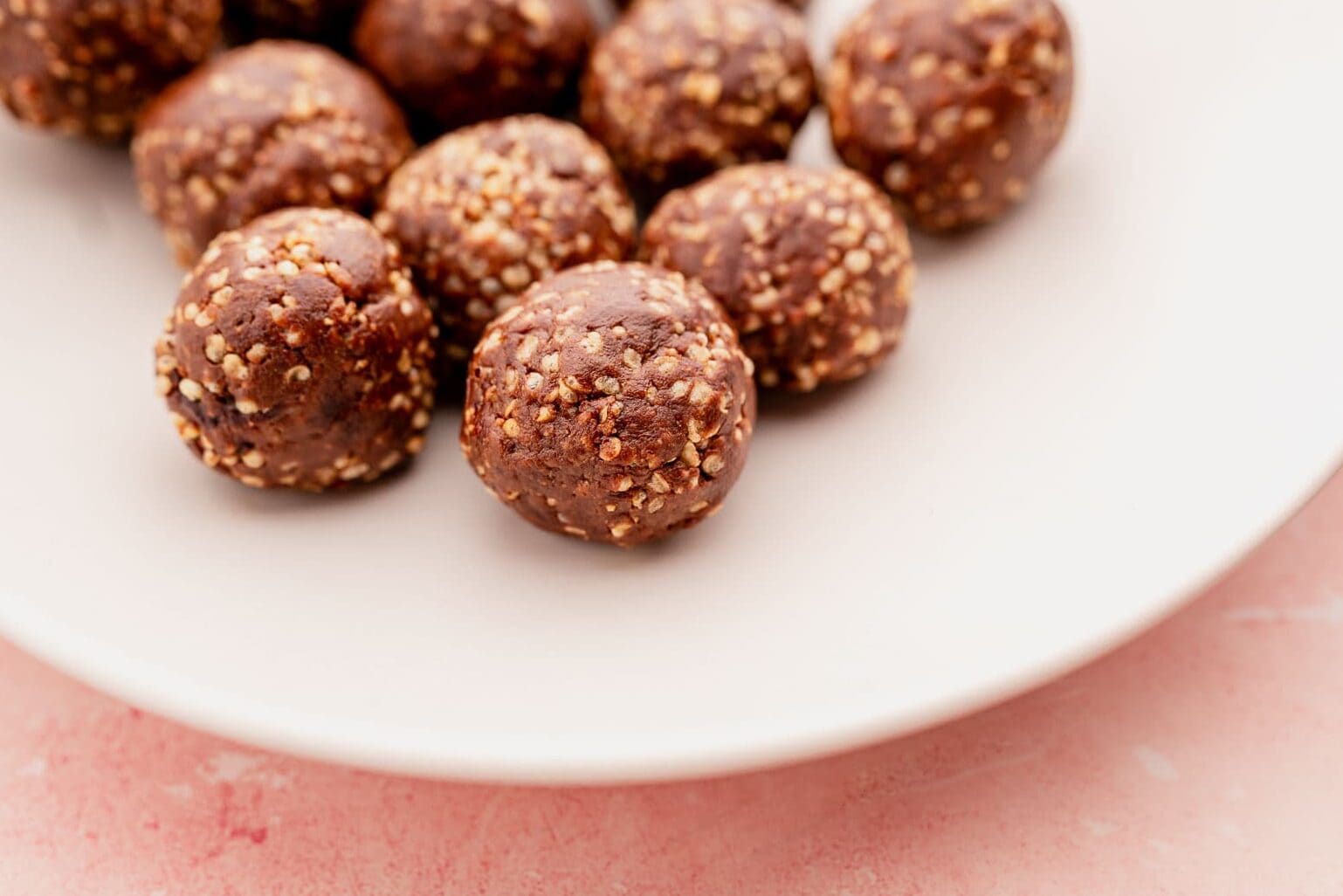 A white plate containing several round, chocolate-covered amaranth seed balls on a light pink surface.