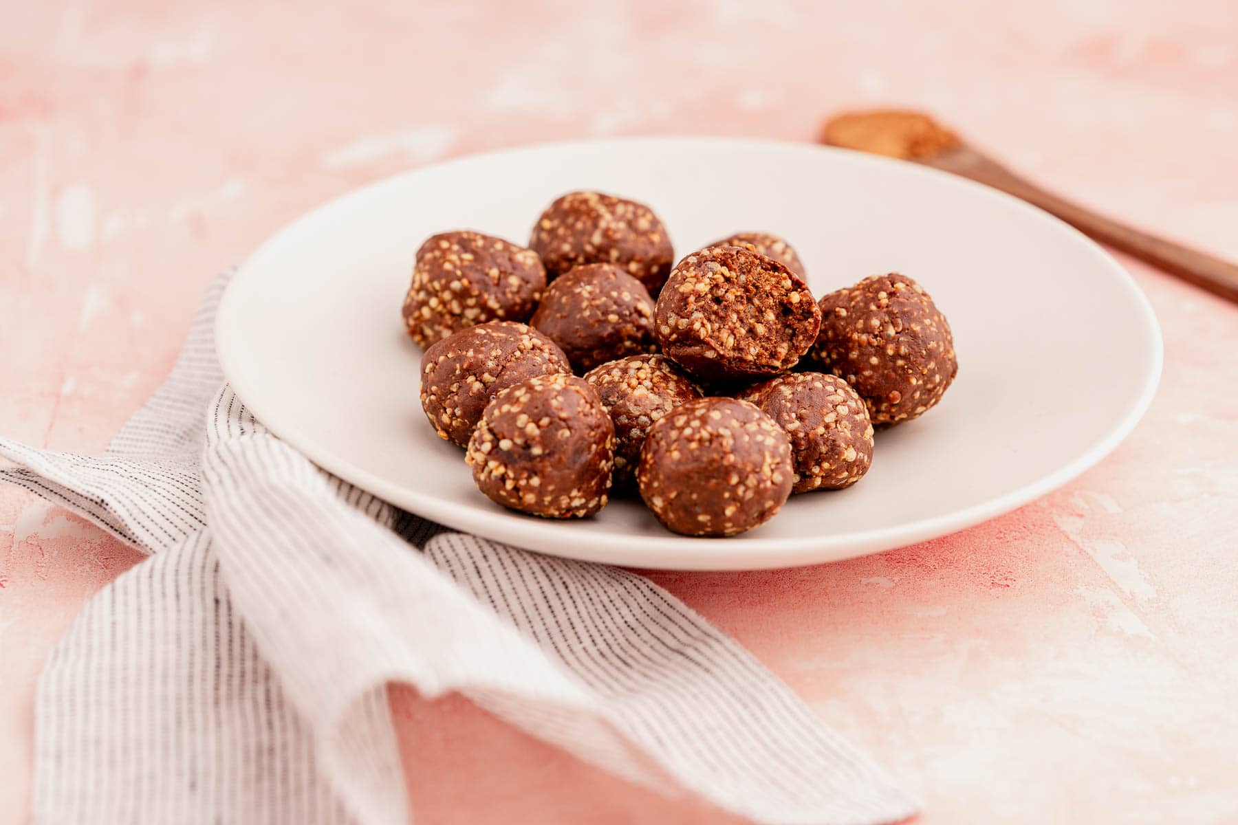 A white plate with chocolate quinoa crunch bites on a pink surface next to a wooden spoon.