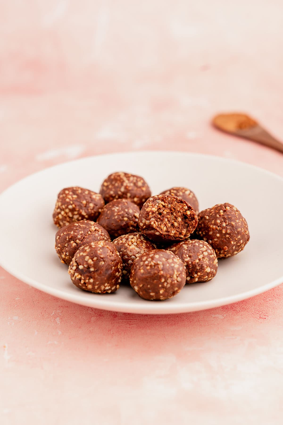 A white plate with homemade quinoa crunch bites sprinkled with seeds, on a pink surface with a wooden spoon in the background.