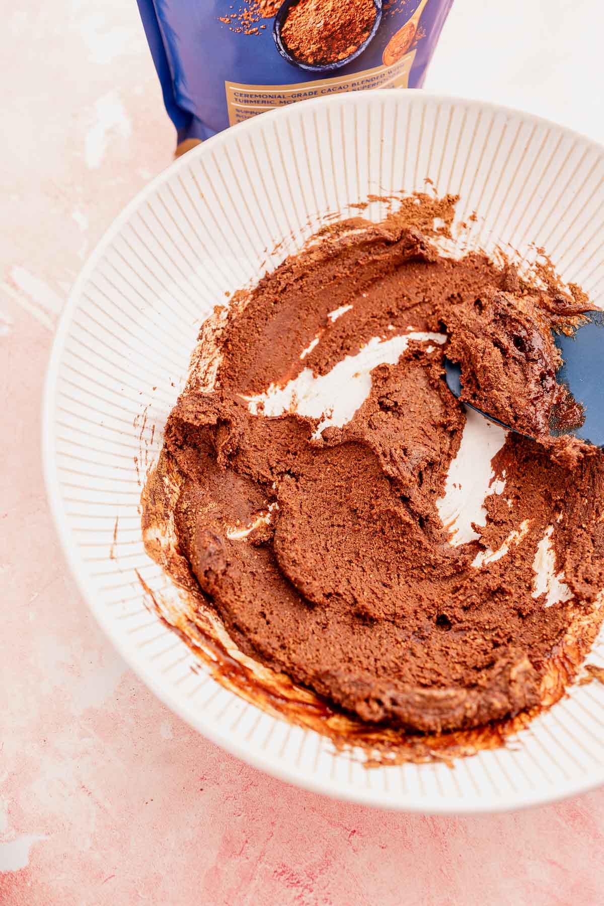 A bowl containing a chocolate and quinoa crunch bites mixture with a spatula, next to an open packet of cocoa powder on a pink surface.
