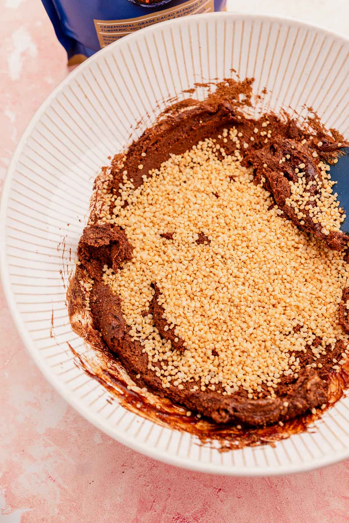A bowl containing a chocolate mixture with quinoa crunch bites sprinkled on top, resting on a pink surface, with a blue container partially visible.