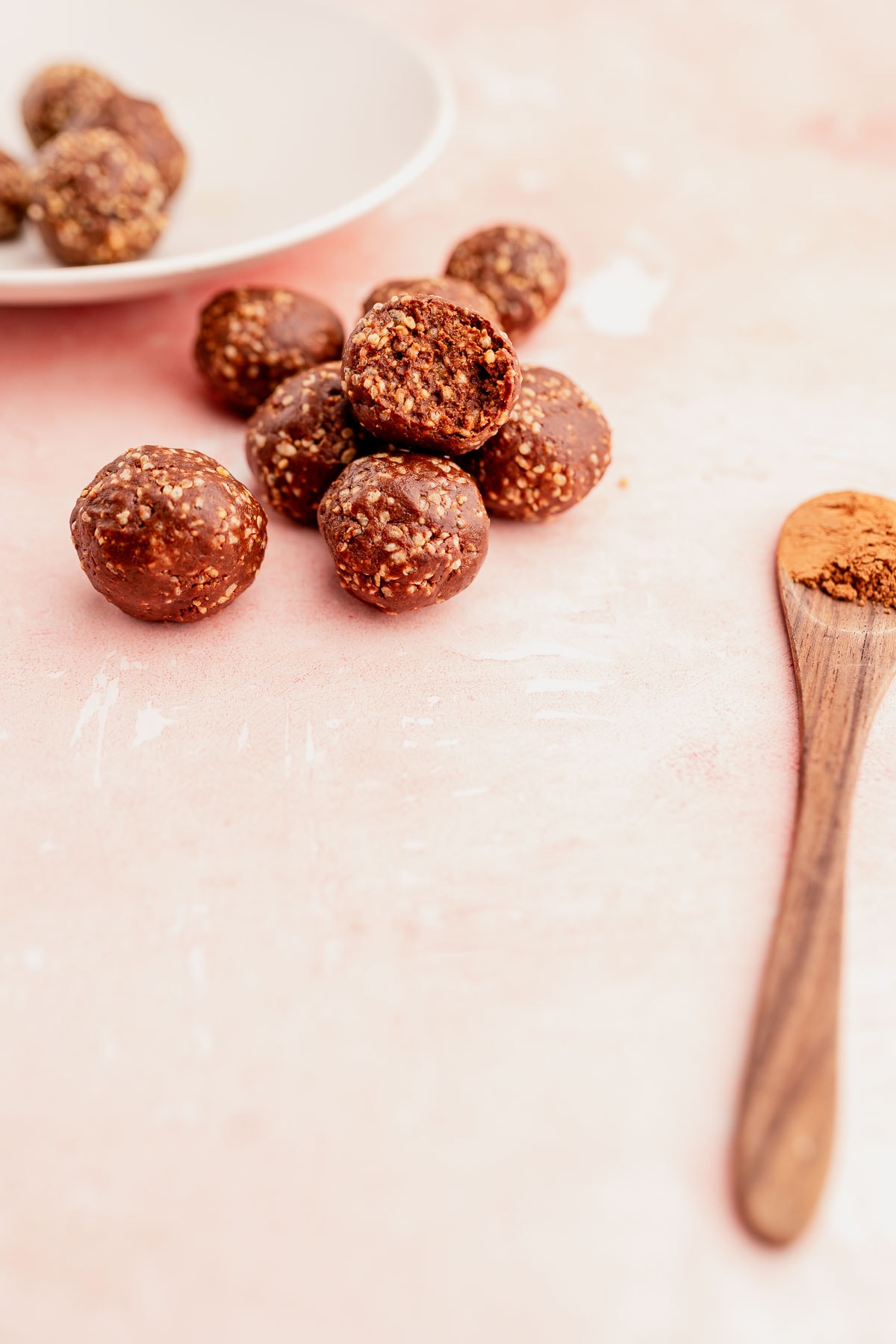 Homemade quinoa crunch bites on a pink surface with some on a white plate and a wooden spoon with cocoa powder.
