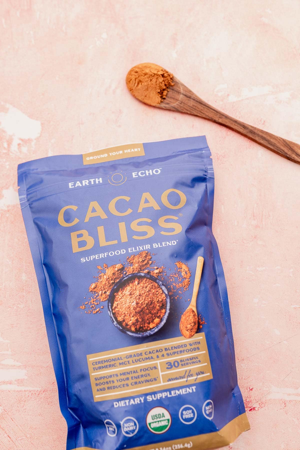 A bag of earth echo cacao bliss superfood elixir blend on a pink surface with a wooden spoon of quinoa crunch bites next to it.