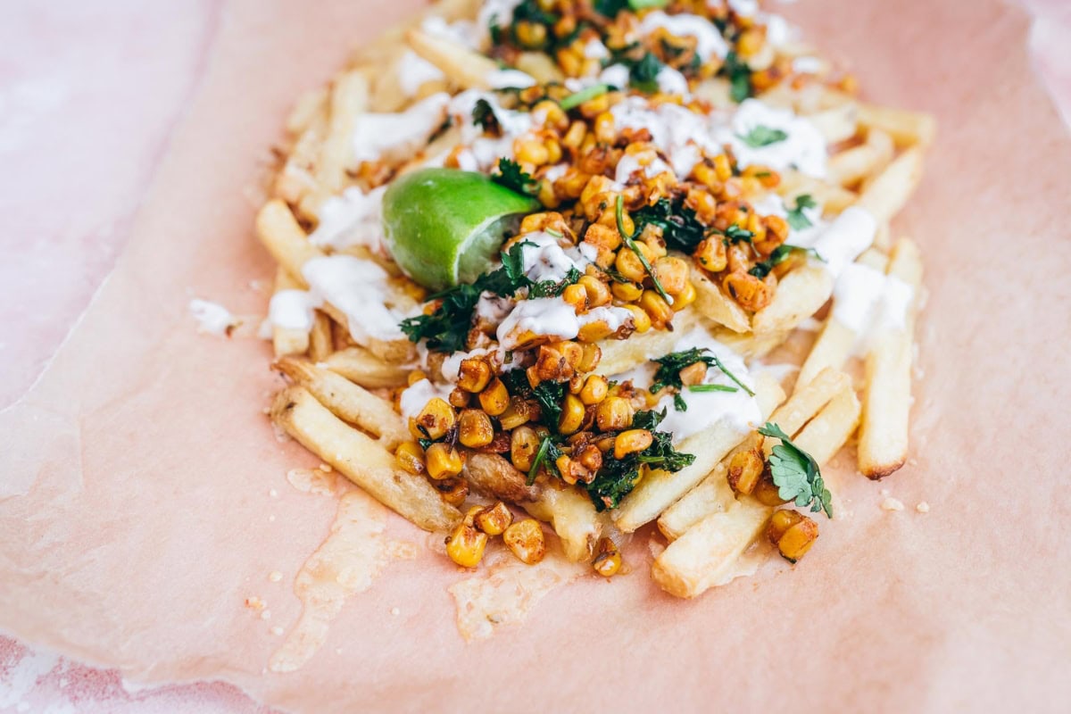 A plate of Mexican-inspired french fries topped with roasted corn, cilantro, a drizzle of creamy sauce, and a lime wedge.
