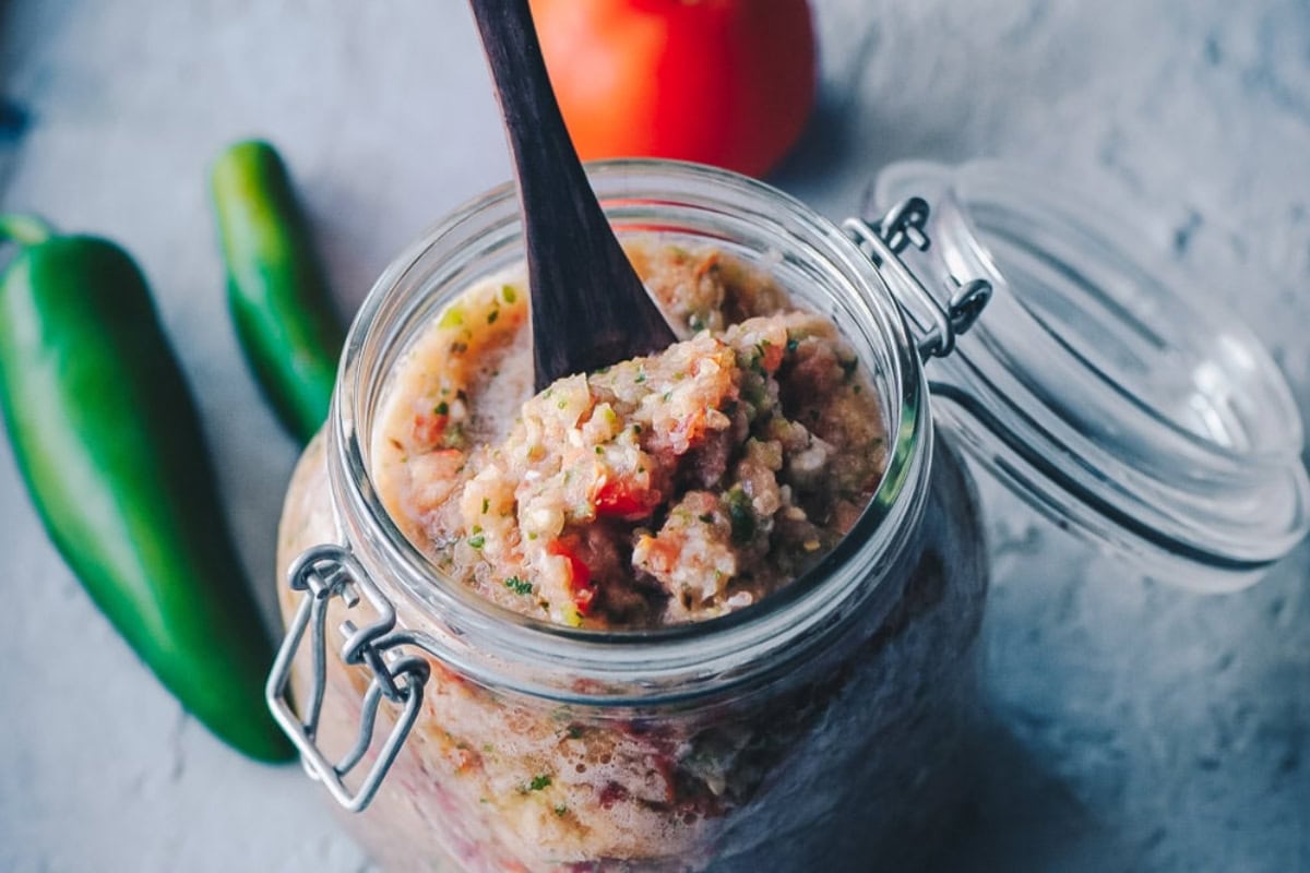 A jar of fresh homemade salsa, inspired by Mexican recipes, with a spoon inside, surrounded by whole tomatoes and jalapeño peppers on a textured surface.