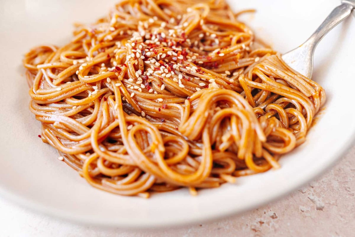 A plate of spaghetti with a sesame seed garnish, coated in a glossy sauce, served with a fork on the side.
