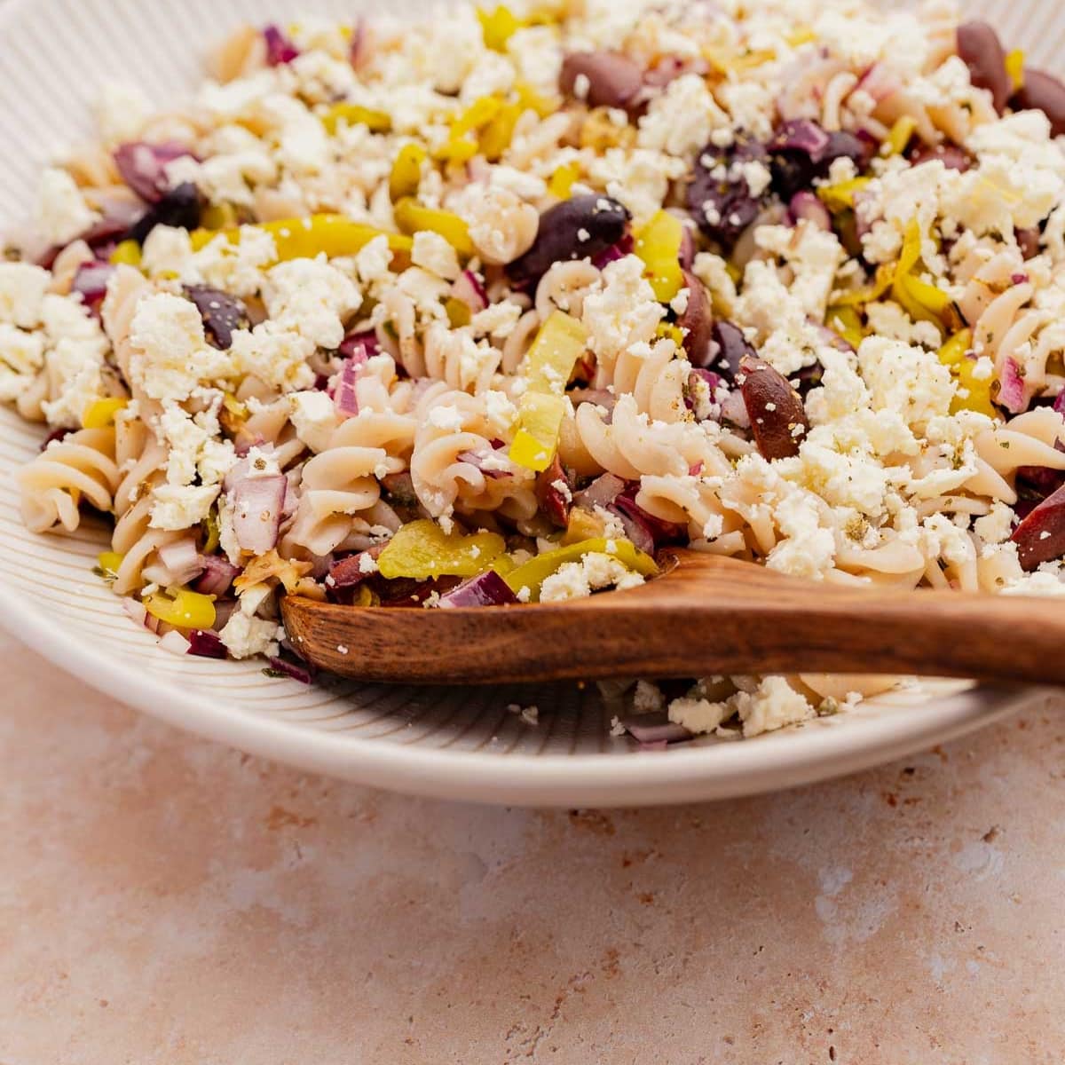 A bowl of Greek pasta salad with black olives, red onions, yellow peppers, and crumbled feta cheese, mixed with a wooden spoon.