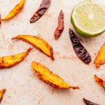 Baked sweet potato wedges and dried mango slices scattered around a halved lime on a light textured surface.