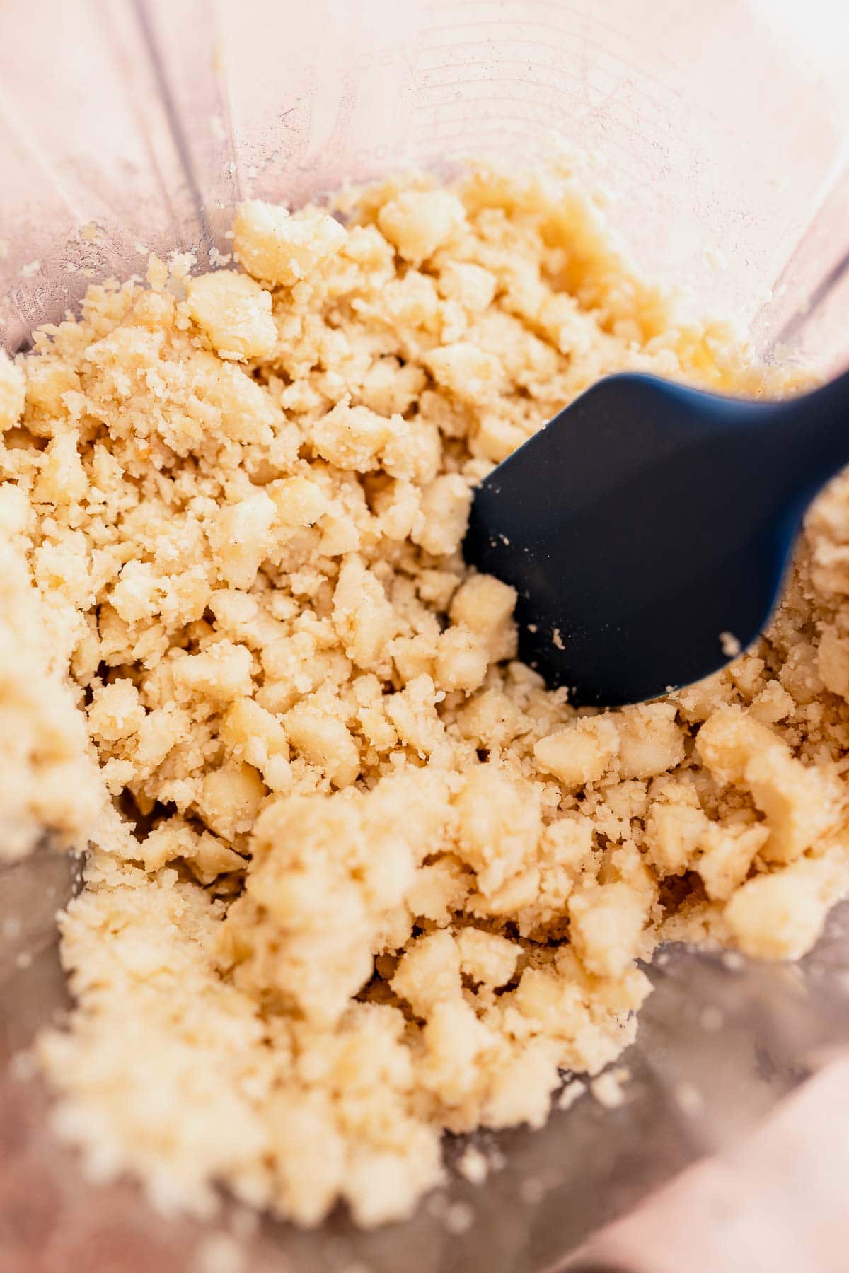 Close-up of crumbled dough with macadamia nut butter in a mixing bowl with a black spatula.