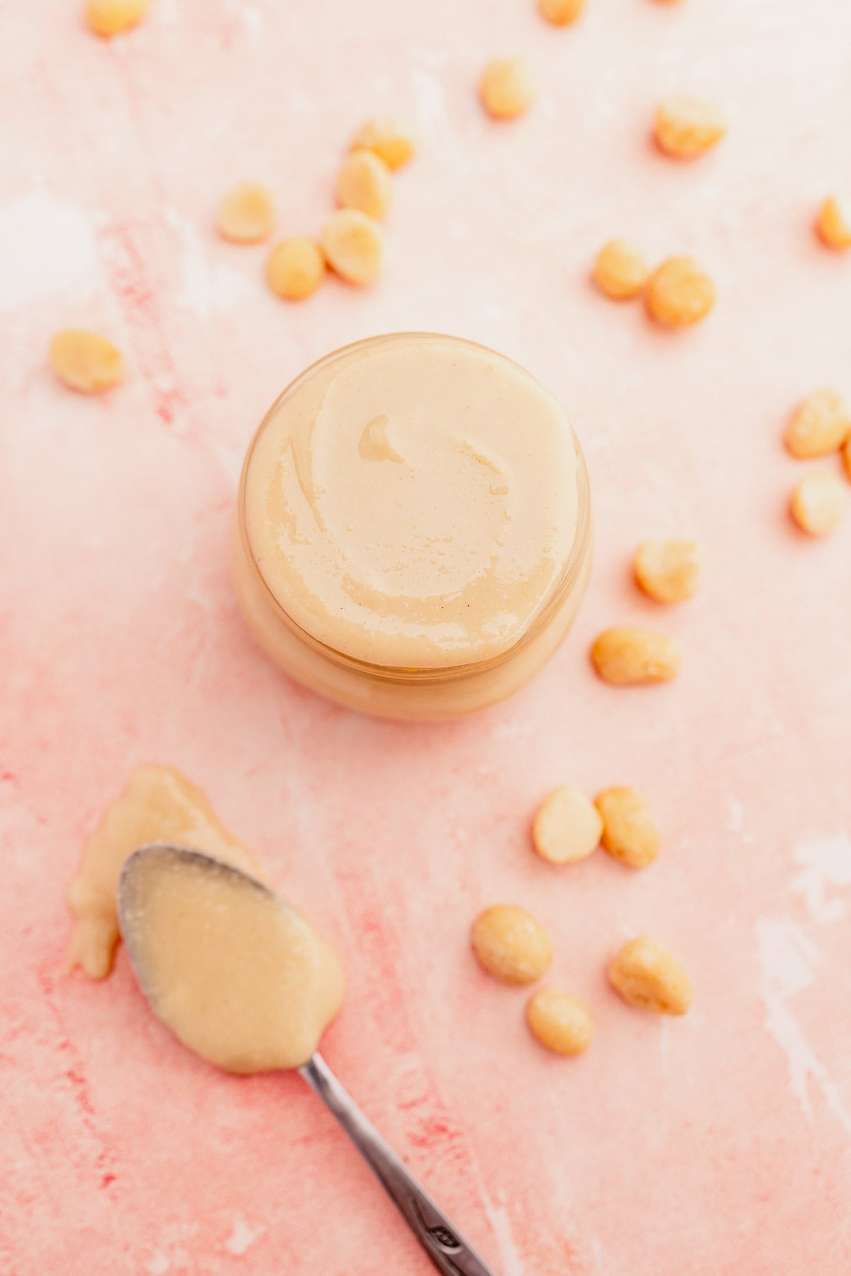 Glass jar of creamy macadamia nut butter with a spoon and spilt nuts on a pink surface.