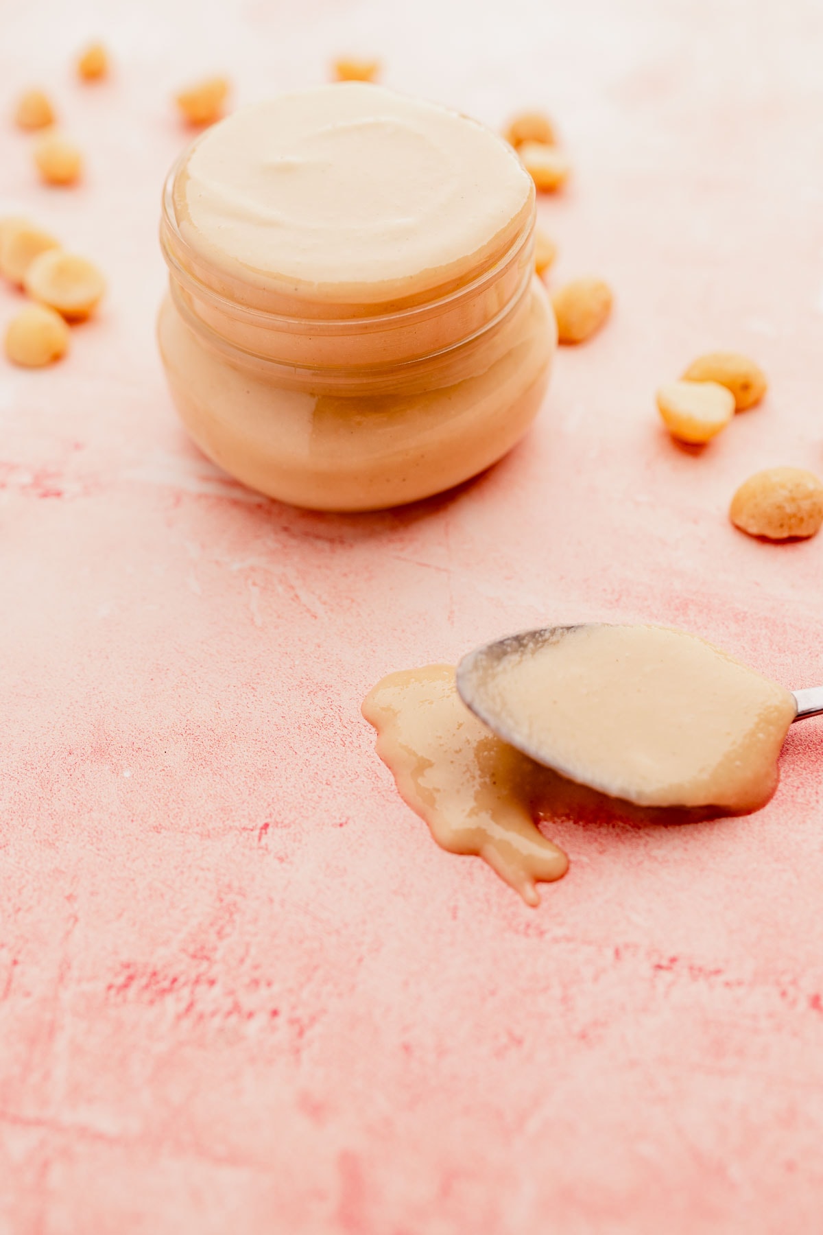 A jar of creamy macadamia nut butter with a spoon, some macadamia nut butter spilled on the tabletop, and scattered peanuts around, on a pink surface.