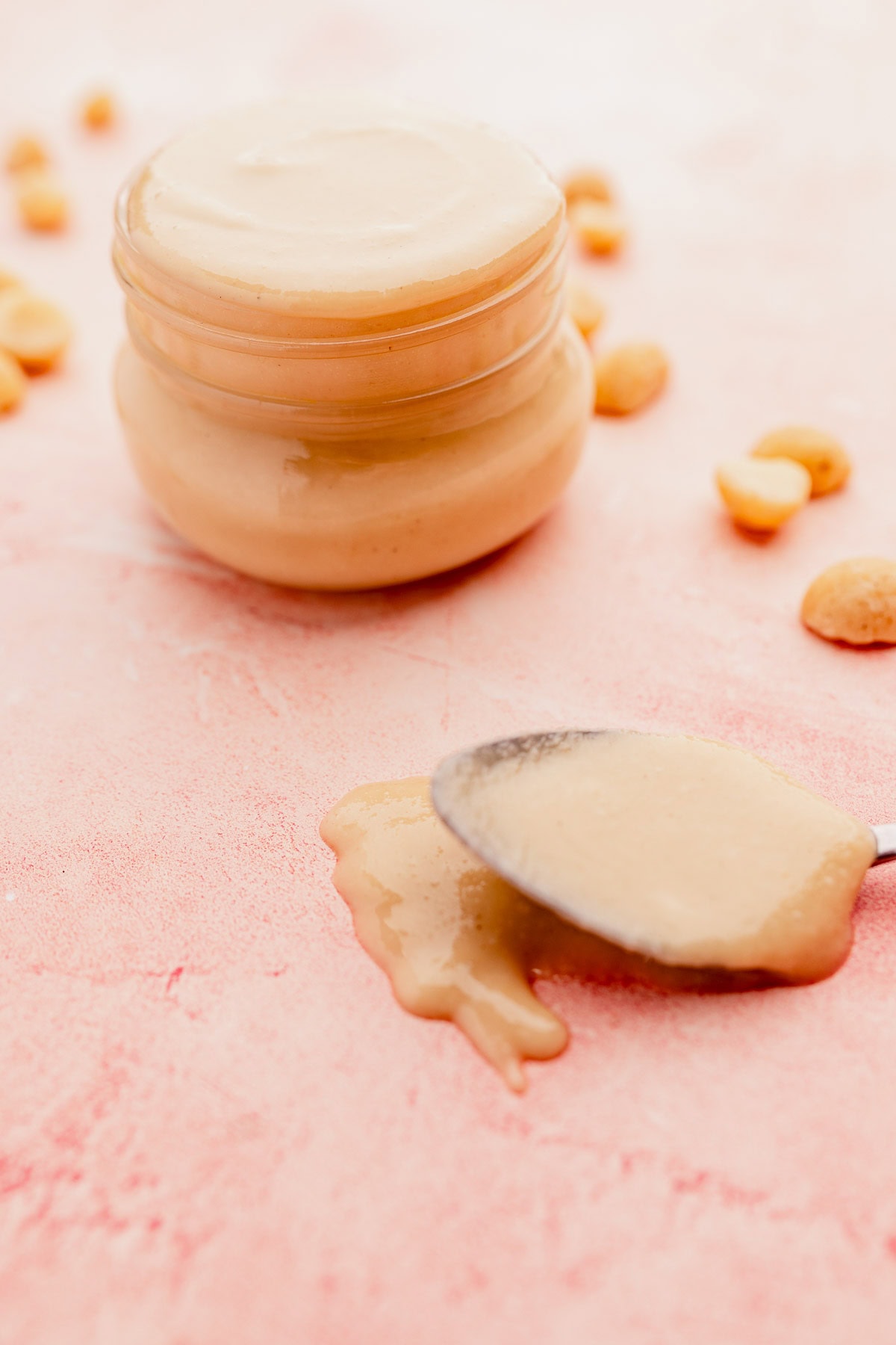 A jar of creamy macadamia nut butter with an open lid and a spoon with nut butter spilling onto a pink surface, scattered nuts around.