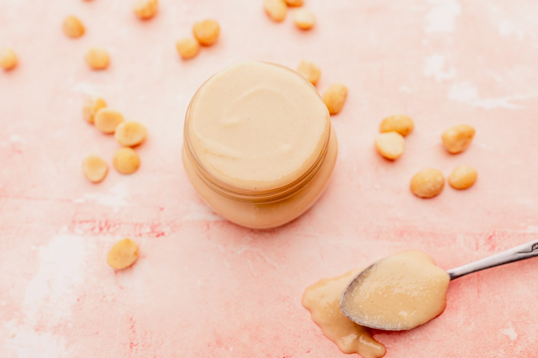 Jar of smooth macadamia nut butter with a spoon beside it, and scattered peanuts on a pink surface.