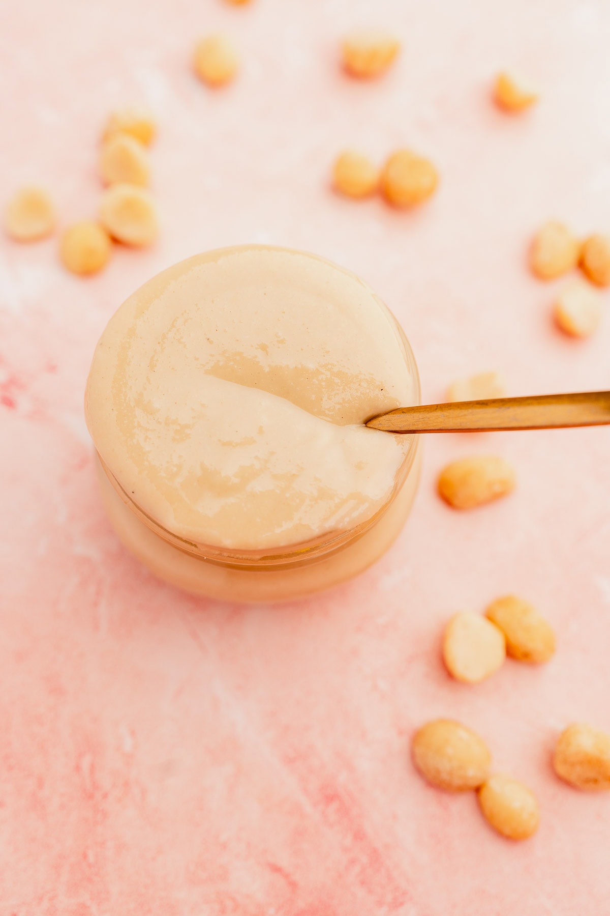 A jar of smooth macadamia nut butter with a wooden spoon on a pink surface, surrounded by scattered peanuts.