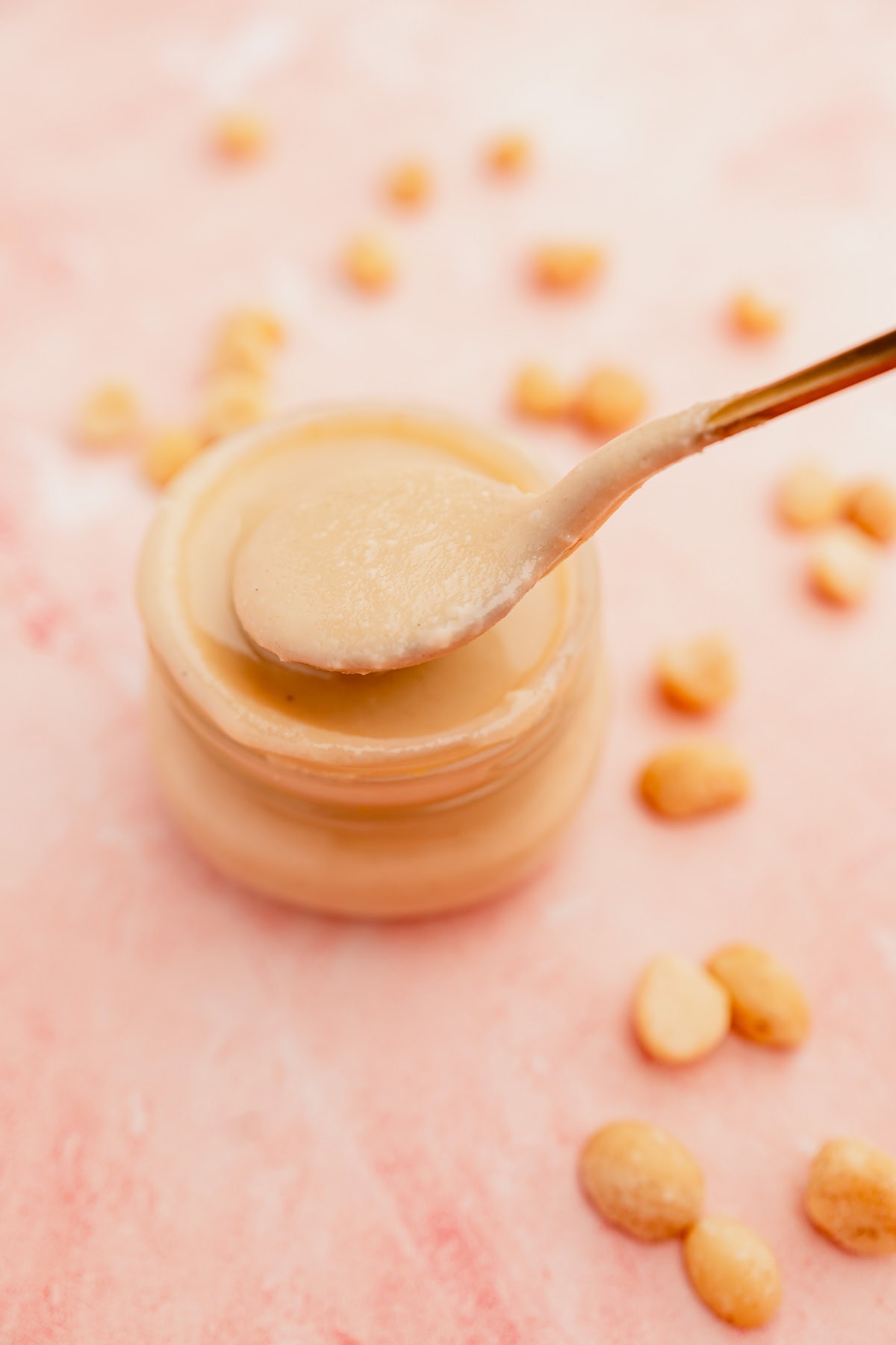 A spoonful of macadamia nut butter over an open jar, with scattered peanuts on a pink background.