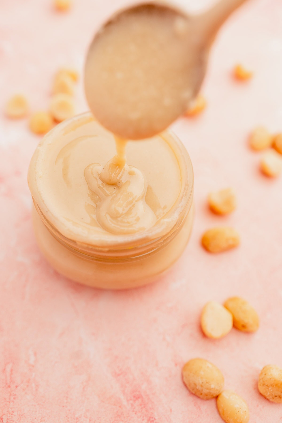 A spoon pours smooth macadamia nut butter into a jar surrounded by whole peanuts on a pink surface.