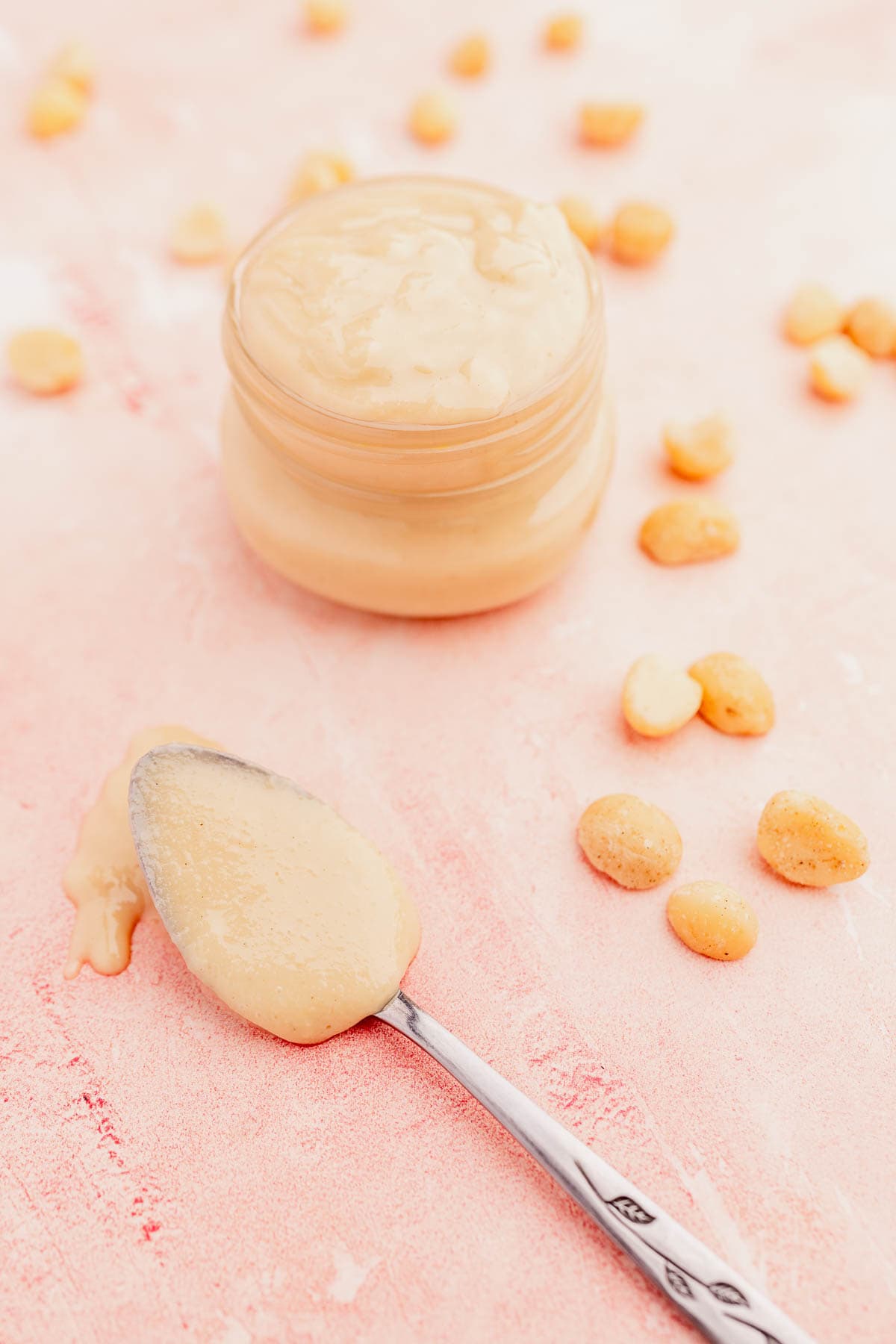 A jar of creamy macadamia nut butter with a spoonful beside it and scattered peanuts on a pink surface.