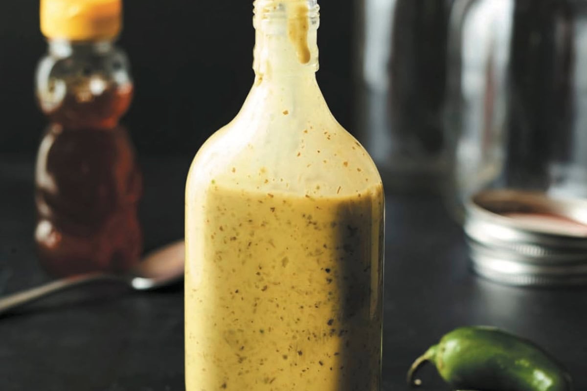 A bottle of creamy, herb-infused salad dressing with visible spice flecks, set against a dark background with a blurred honey jar in the background.