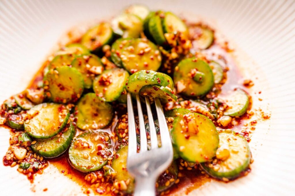 A close-up image of a dish with sliced cucumbers, chili flakes, and sesame seeds, garnished with a fork on the side.