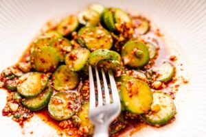 A close-up image of a dish with sliced cucumbers, chili flakes, and sesame seeds, garnished with a fork on the side.