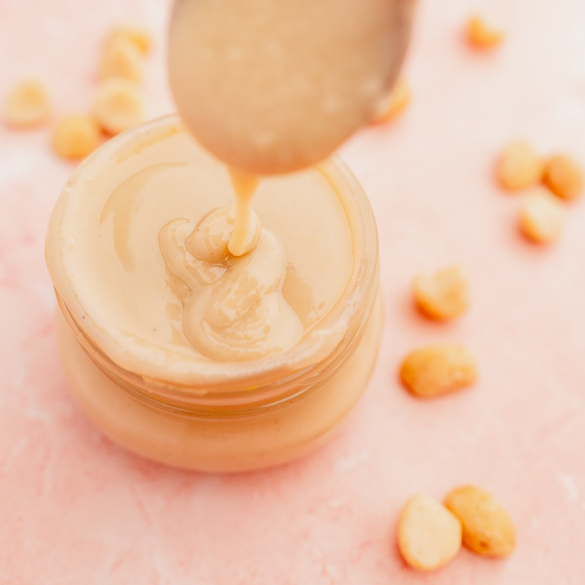 A spoon driops creamy macadamia nut butter into a glass jar on a pink surface, surrounded by scattered peanuts.
