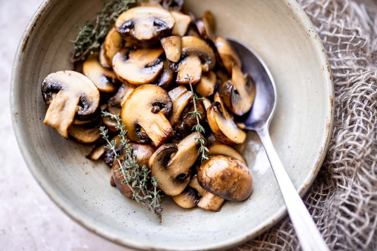 A bowl of sautéed mushrooms garnished with thyme, served with a spoon on a textured beige cloth.
