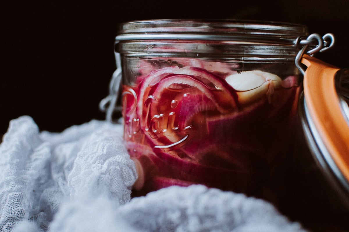 A glass jar filled with pickled red onions and garlic cloves, partially opened and set on a white lace cloth against a dark background.
