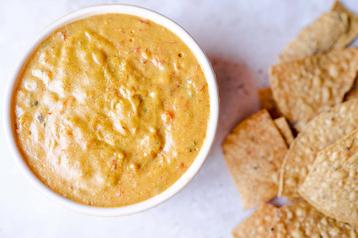 A bowl of creamy, orange-colored queso dip with diced vegetables, accompanied by tortilla chips on a marble surface.