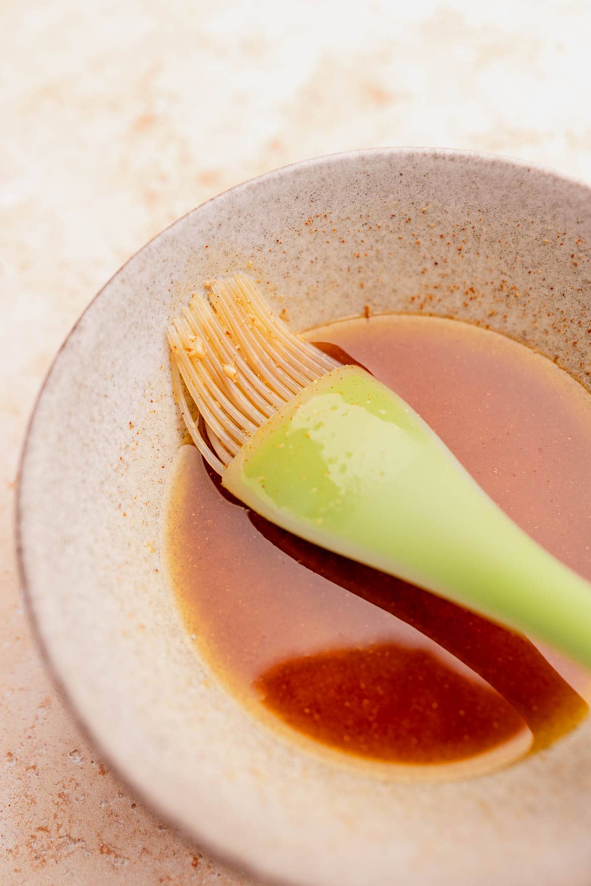 A ceramic bowl with soy sauce and a green brush lying across it on a textured surface with cabbage steaks.
