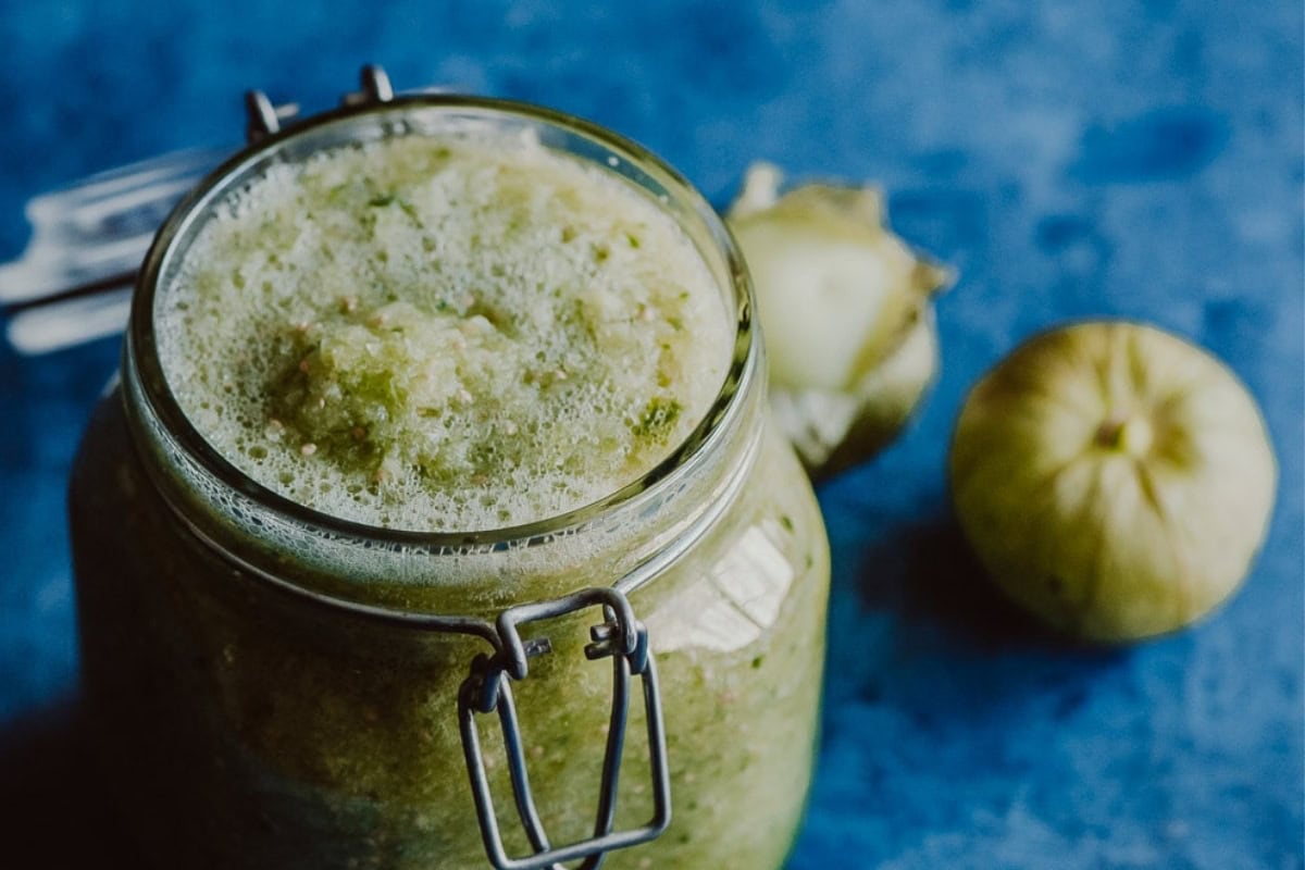 A jar of fresh green tomatillo salsa sits beside whole tomatillos on a blue textured surface.